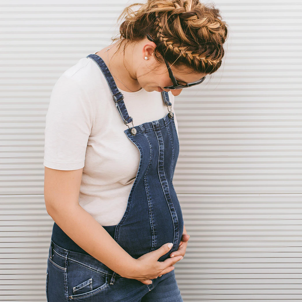 A pregnant woman in denim overalls gently holding her stomach, representing healthy pregnancy.