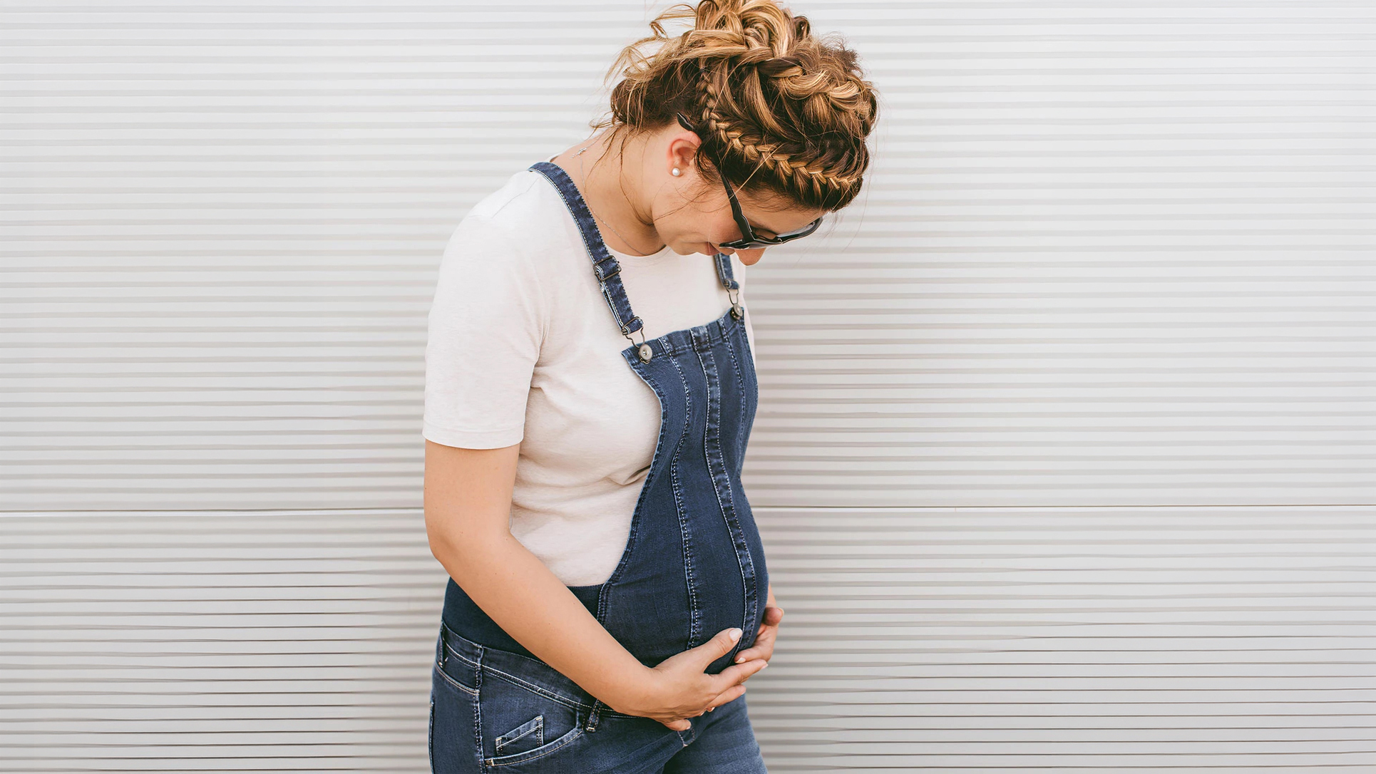 A pregnant woman in denim overalls gently holding her stomach, representing healthy pregnancy.