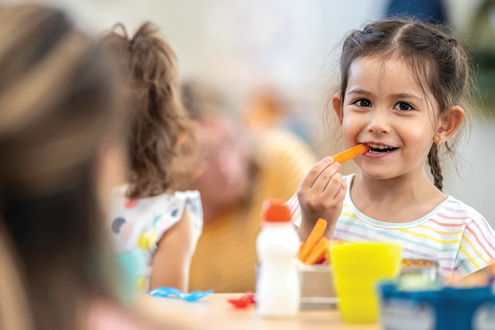 Girl eating lunch, smiling while holding carrot stick